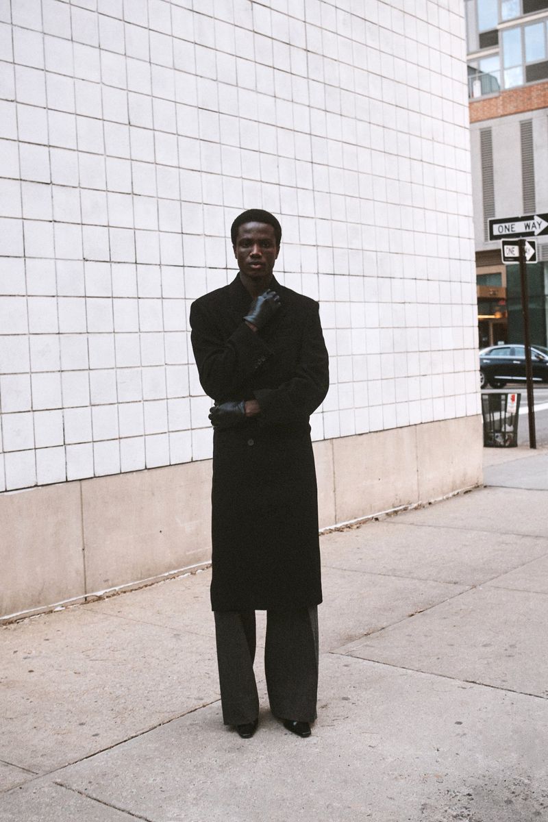 A full shot of a stylish black man in a long coat and gloves, posing on a city sidewalk with a tiled building in the background.