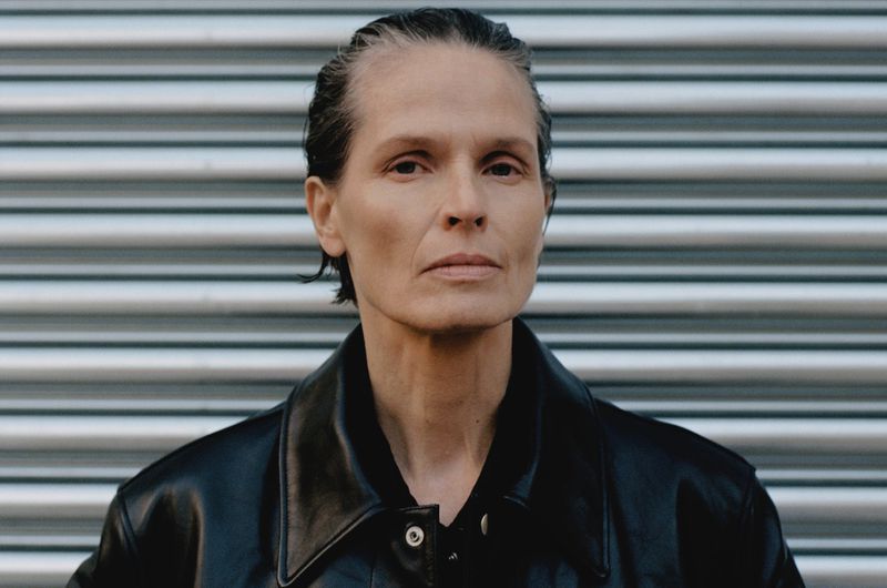 Close-up studio portrait of an adult female with short, greying hair wearing a dark leather jacket against a gray metal background.