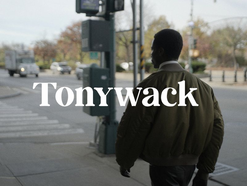 A man in a bomber jacket walking on a sidewalk. Vehicles and greenery are visible in the background on an overcast day.