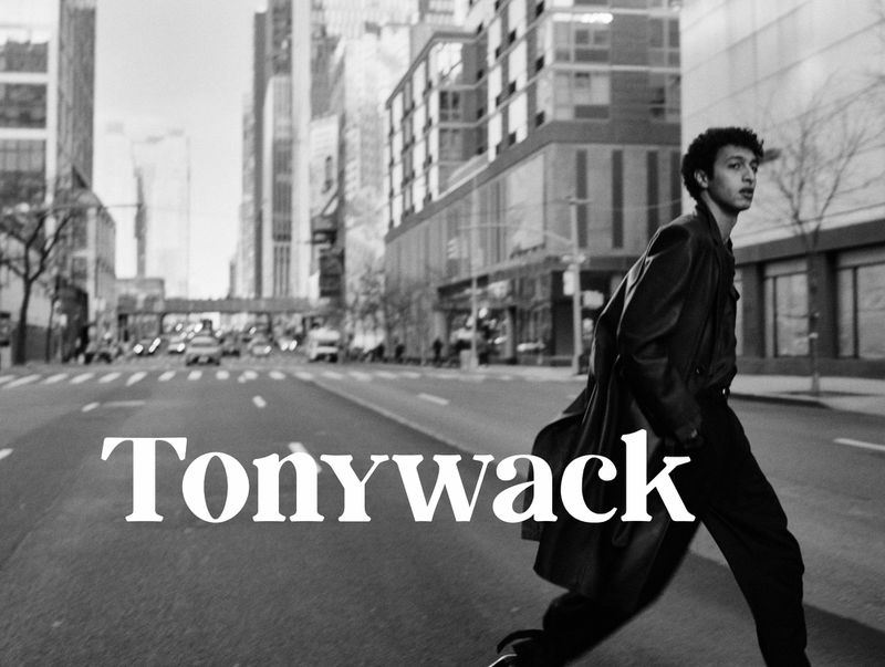 Monochrome shot of a young adult male confidently walking across a city street with skyscrapers, wearing a dark coat and carrying a bag.