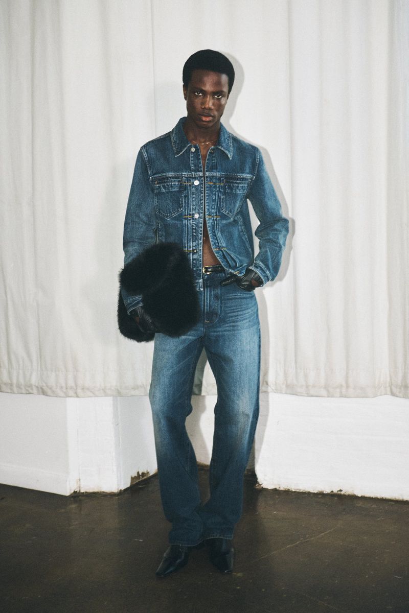Full shot of a Black male model wearing a denim jacket, jeans, and black leather gloves while holding a furry bag in a studio setting.