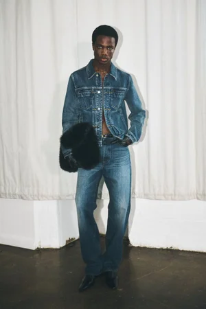 Model in denim jacket and matching jeans holding a dark furry bag standing before draped white fabric