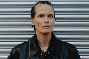 Close-up portrait of a model with slicked-back dark hair wearing a black leather jacket against a corrugated metal shutter background.