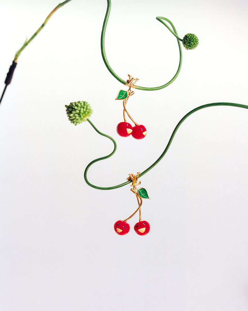 Two cherry pendants hang from green vines on a white background, showcasing a playful and whimsical design in a minimalist composition.