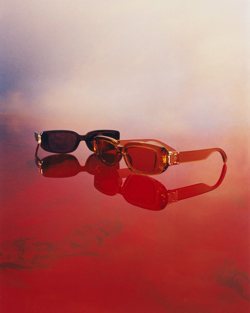 Two pairs of sunglasses, one amber and one dark brown, are displayed on a shiny red surface with a bright gradient background.