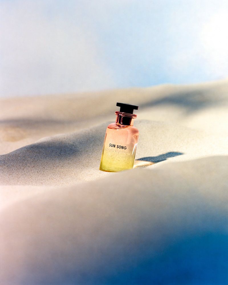 Elevated shot of a perfume bottle on a sandy surface, bathed in natural light with a blue sky visible in the background.