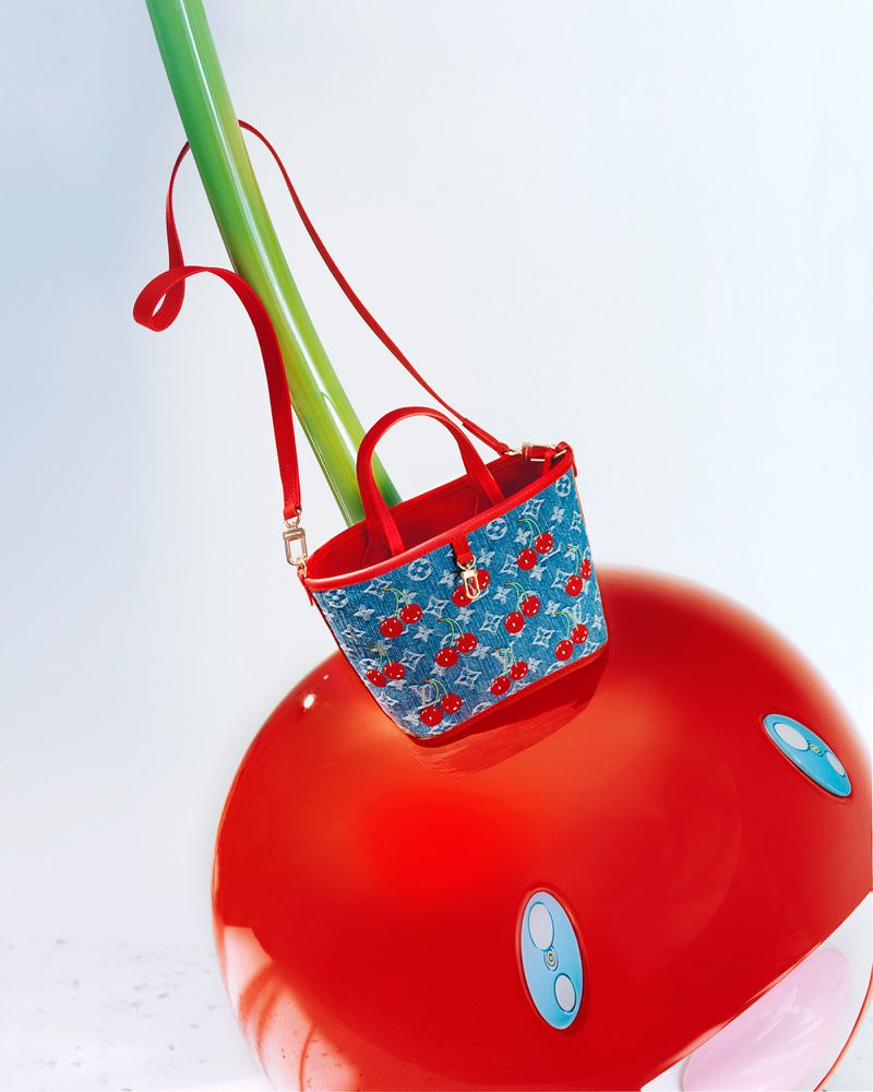 Designer handbag with cherry pattern displayed on a large cherry prop against a clean, white background in a studio setting.