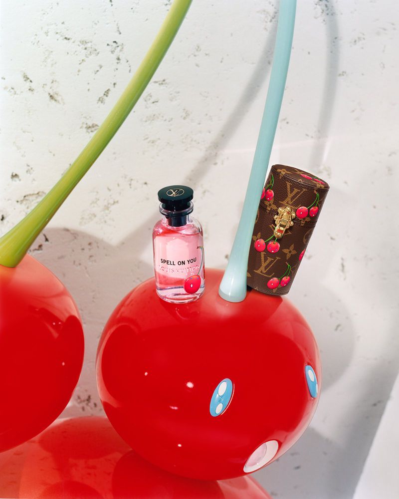 Louis Vuitton perfume bottle and cherry print case sit atop a red decorative cherry with a light grey marbled background.