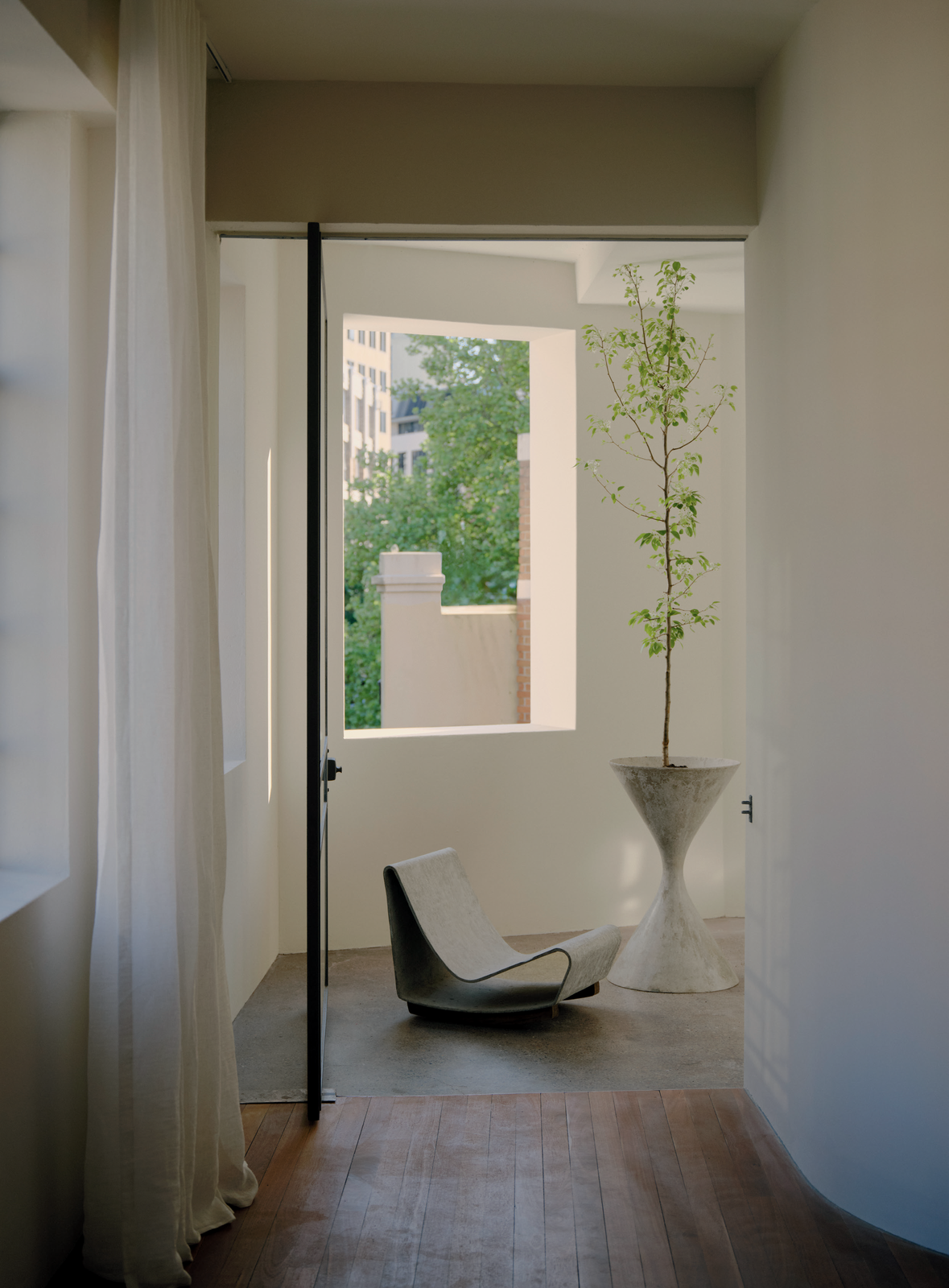 Indoor interior featuring a modern chair, a potted tree on a pedestal, hardwood floors, and natural light from a window.