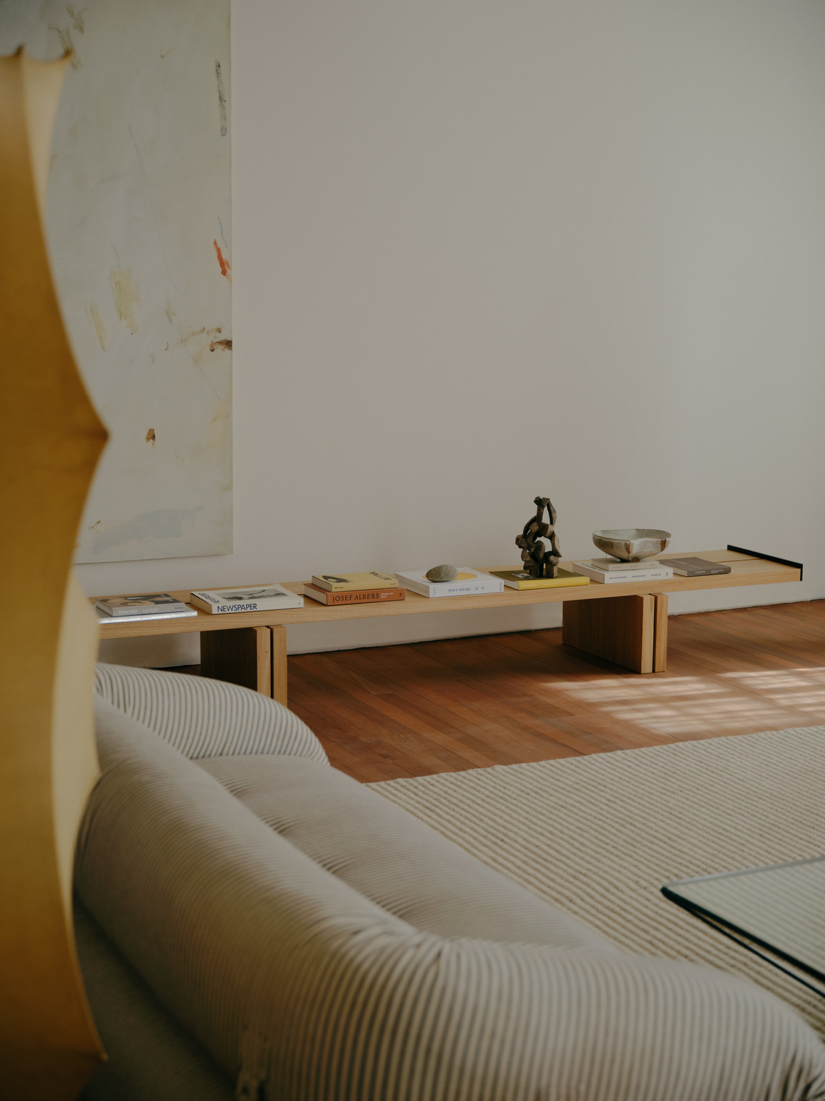 Living room interior with a beige striped couch, a light wood shelf holding books and decorative objects against a white wall, and a wooden floor.