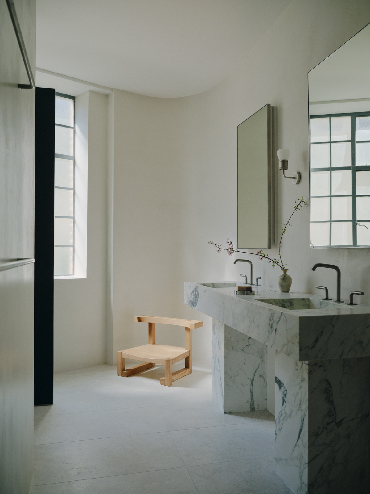 Minimalist bathroom with marble double vanity, light wood chair, and natural light from a large window. Clean design, luxury home.