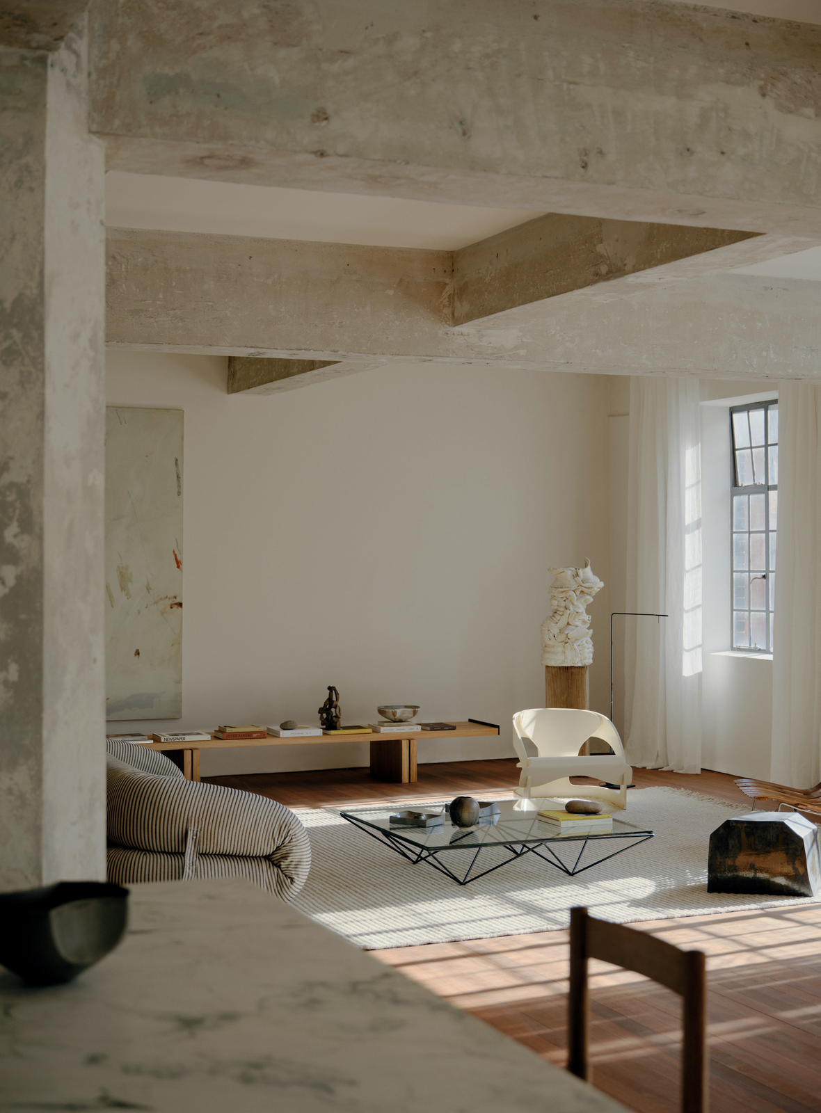 Sunlit living room interior with modern furniture, including a glass coffee table, a minimalist sofa, and a sculptural art piece.