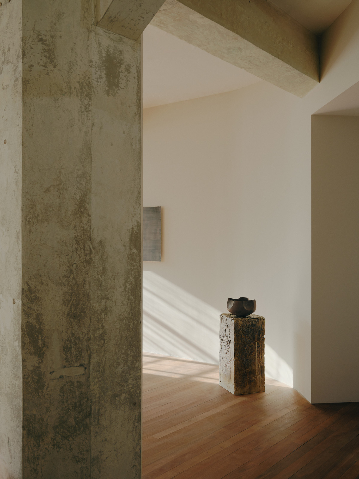 Minimalist interior featuring a dark bowl on a concrete pedestal, framed by architectural concrete beams and light wood flooring.