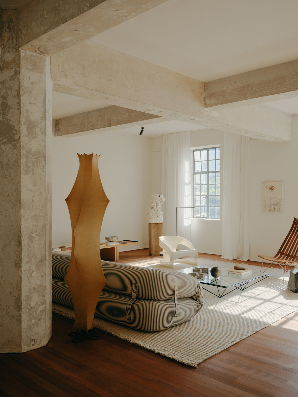 Minimalist living room interior with a low striped sofa, sculptural floor lamp, and large window with white curtains.
