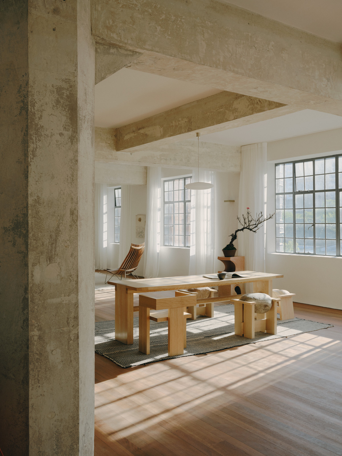 Minimalist interior of an open loft space featuring a wooden dining table with benches and sunlight streaming through large windows.