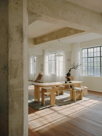 Minimalist interior of an open loft space featuring a wooden dining table with benches and sunlight streaming through large windows.