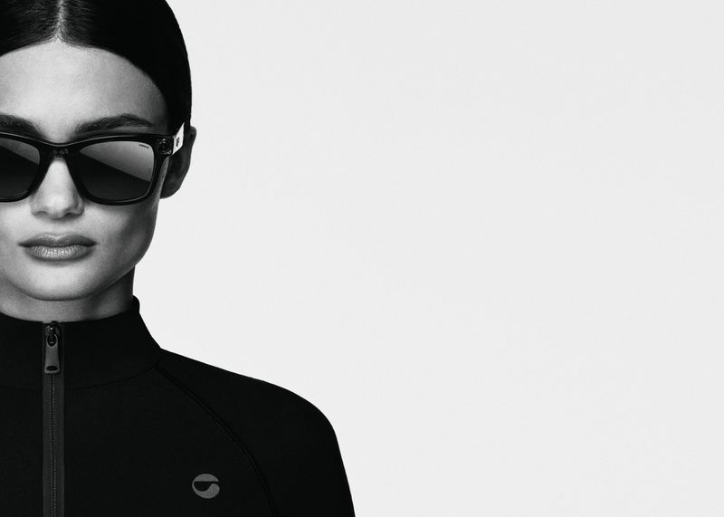 Portrait of a woman wearing black sunglasses and a black zip-up top against a bright white background in a studio setting.