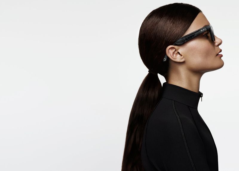 Profile of a woman with dark hair in a ponytail wearing black smart glasses against a bright white background in a studio shot.