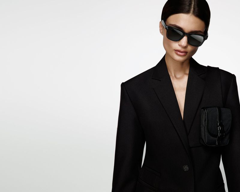 Stylish woman in a black blazer and sunglasses poses against a clean, white backdrop, showcasing a modern and chic fashion aesthetic.