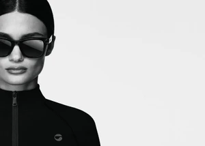 Close-up portrait of a model wearing dark sunglasses and a black zipped top against a solid white background.