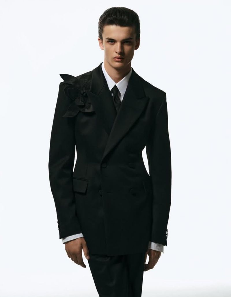 Male model in a black double-breasted tuxedo suit with a floral accent on the shoulder poses against a bright white background in a studio.