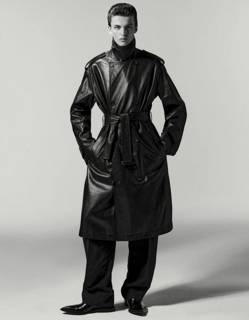 Full-length portrait of a young adult male fashion model in a belted black leather trench coat, dark turtleneck, dress pants, and shiny dress shoes.