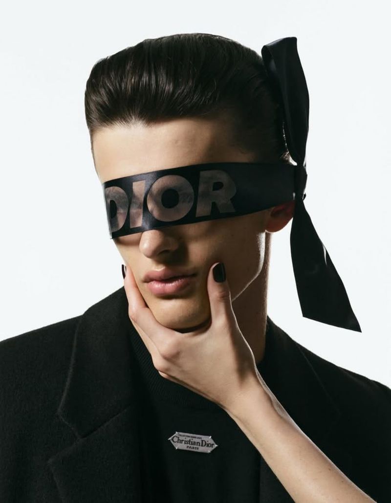 Close-up of a model with dark hair wearing a black blindfold with the Dior logo, juxtaposed with a woman's hand gently touching his face.