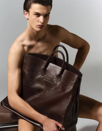 Shirtless male model Tristan Watkins seated against a grey studio backdrop holding a large dark brown leather tote bag.