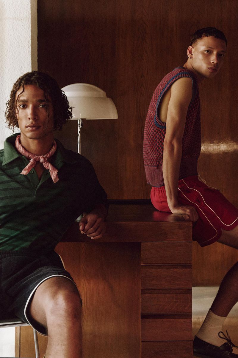 Two young adult men in vintage-inspired fashion, posed indoors with a retro lamp. One man is seated; the other is standing near a wooden desk.