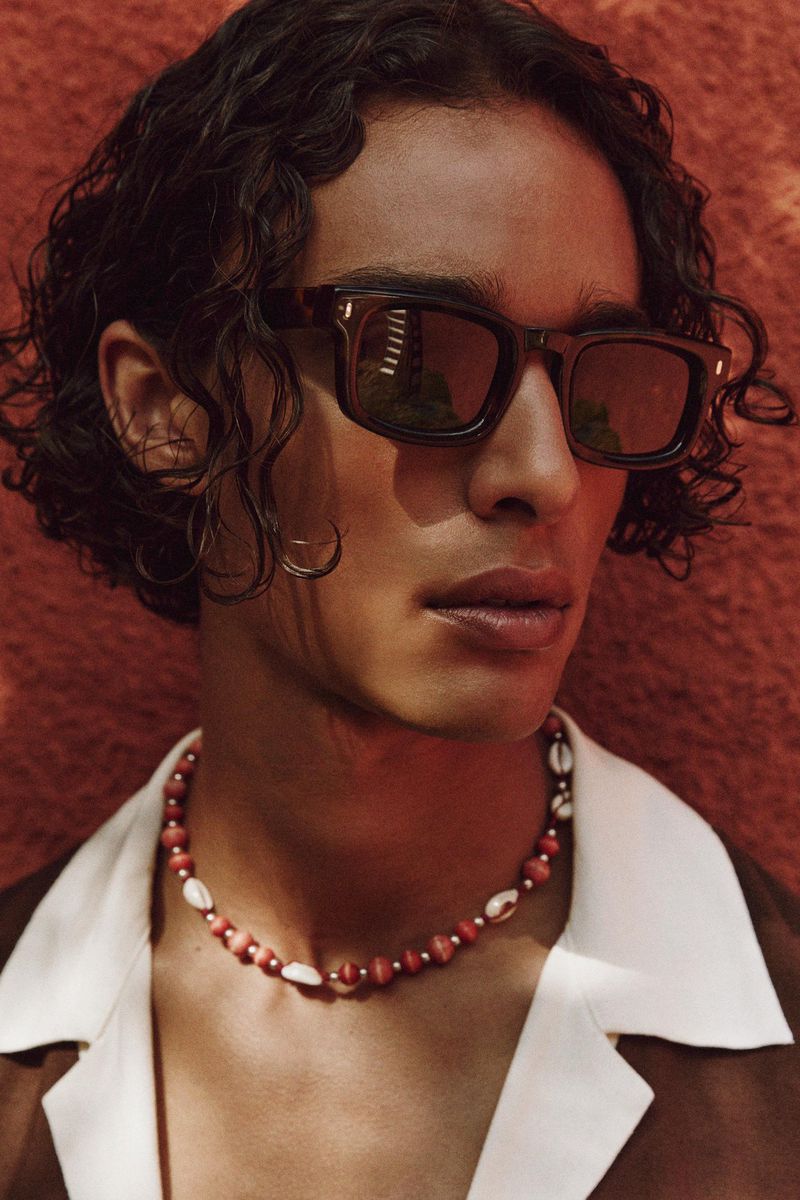 Portrait of a young adult male with curly hair, wearing sunglasses and a beaded necklace, posing against a textured red background.