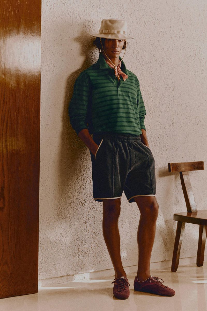 A man with dark hair and a light complexion stands leaning against a cream-colored wall wearing a green shirt, dark shorts, and burgundy sneakers.