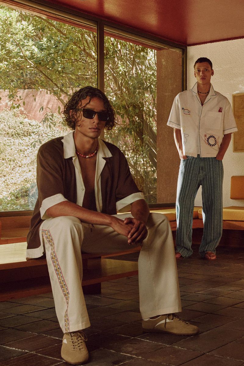 Two adult males pose indoors with natural light streaming through a large window. One is seated, and the other standing in the background.