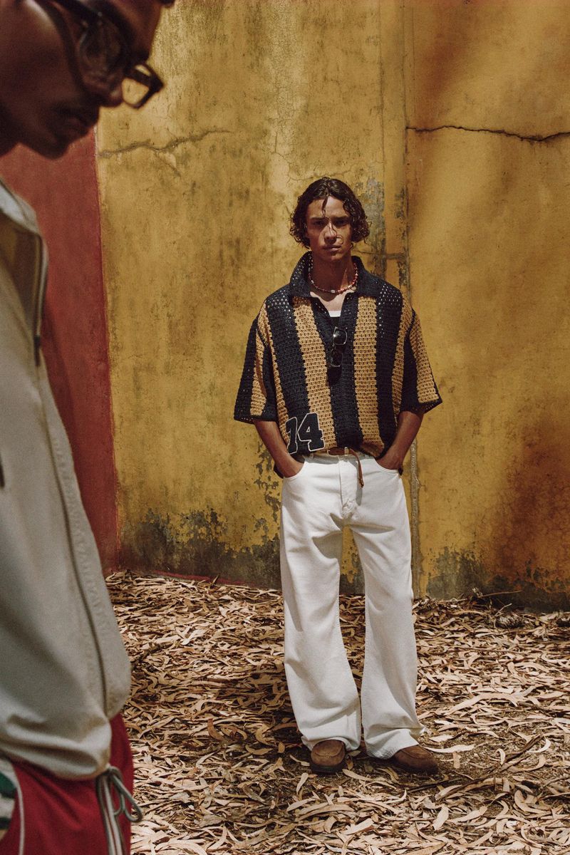 A young man stands in front of an aged wall in a striped shirt and white jeans with another person partially visible on the left.