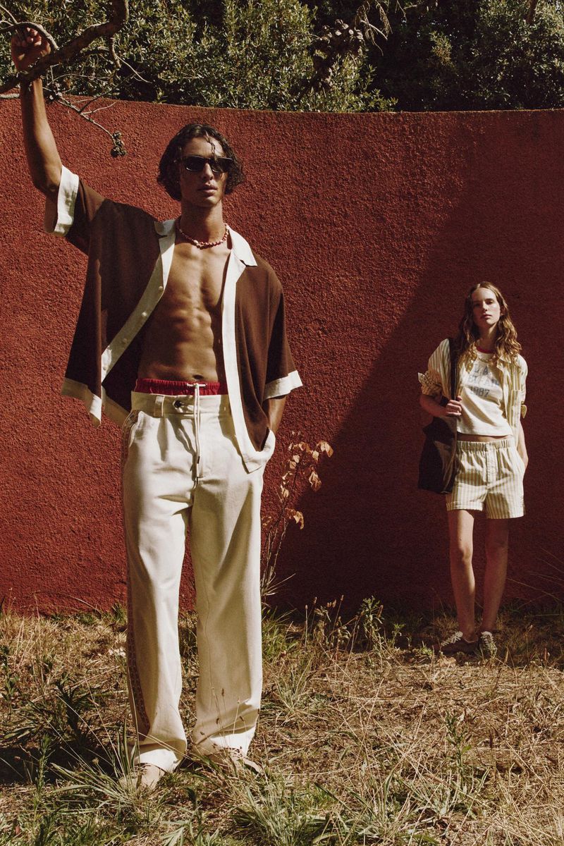 A man and woman pose in outdoor fashion shot; the man wears a brown shirt and cream pants, while the woman wears a beige outfit.