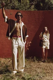 Djairo Mulder in an open brown knit shirt and cream trousers poses by a red textured wall for Zara's The Embroidery Edit.