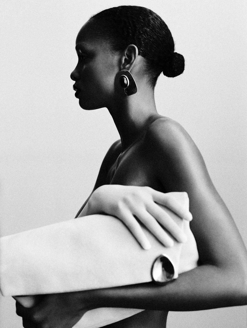 Black and white fashion photo: a stylish Black woman in profile holds a large clutch. Earrings and hair up complement the simple studio backdrop.
