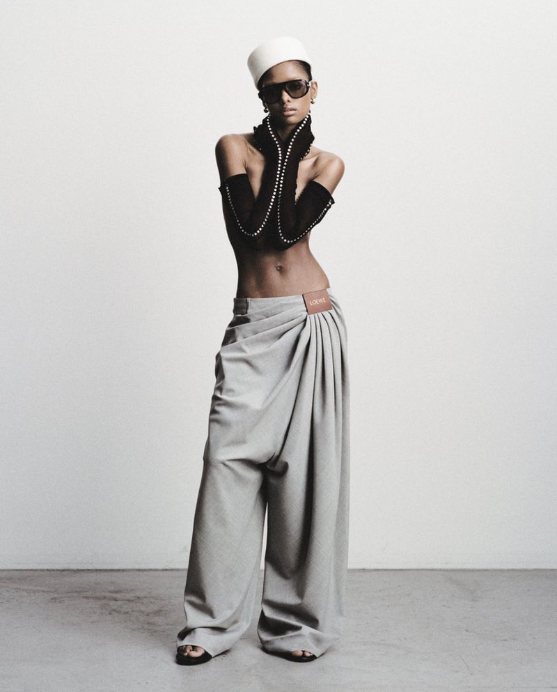 A person models draped grey pants with exposed midriff, wrist-length beaded gloves, and a white hat, against a neutral backdrop in a fashion editorial.