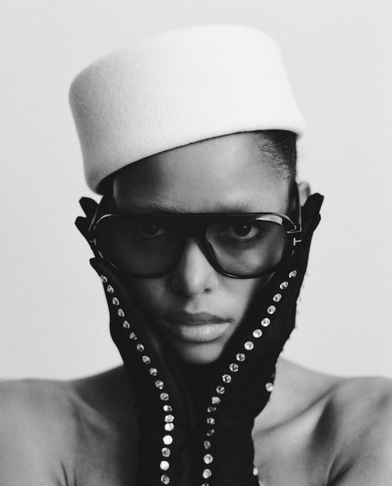 Monochrome portrait of a person wearing a white hat, sunglasses, and gloves, with hands framing their face against a neutral backdrop.