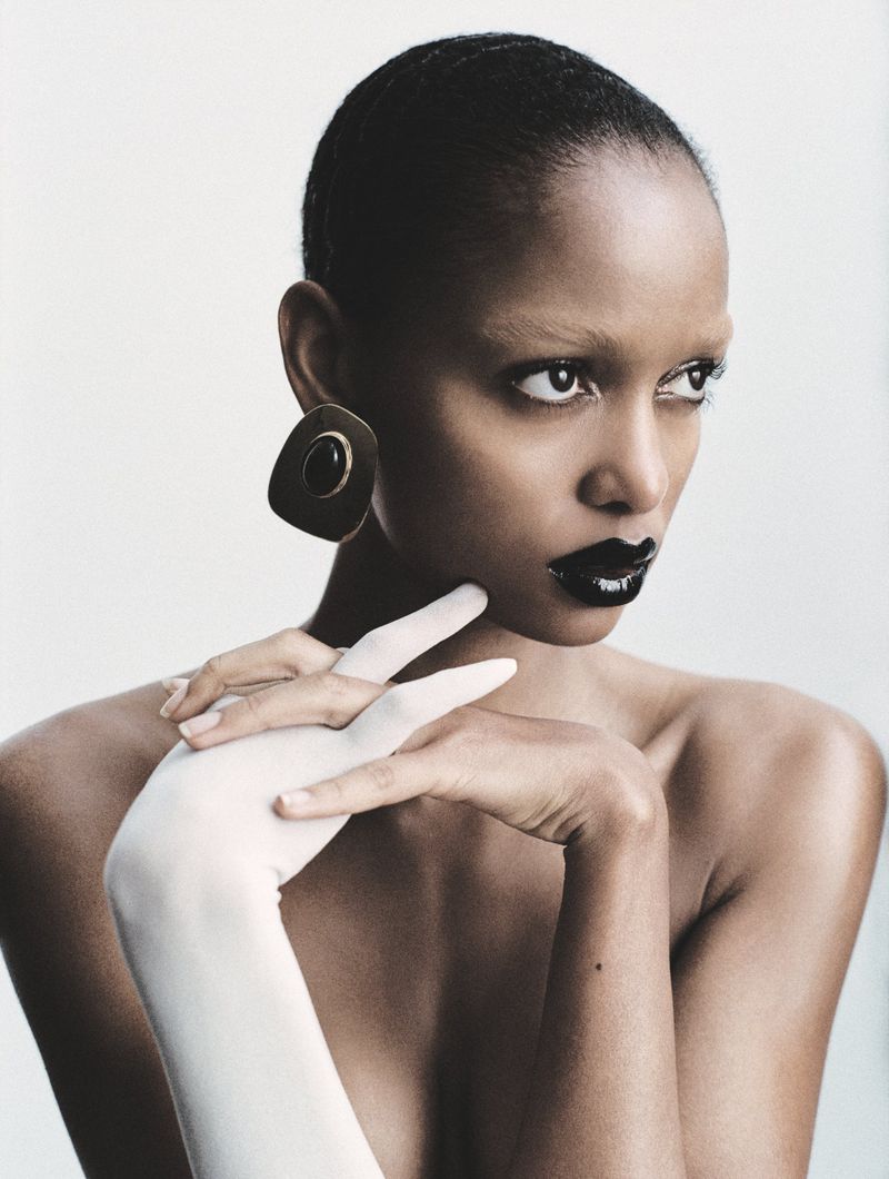 Portrait of a black woman with short hair, wearing an earring and lipstick, posing with her hands and a gloved hand in a photo studio.