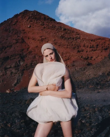Model Julia Banas wearing a sculptural white mini dress and head covering in a rugged red volcanic mountain landscape.