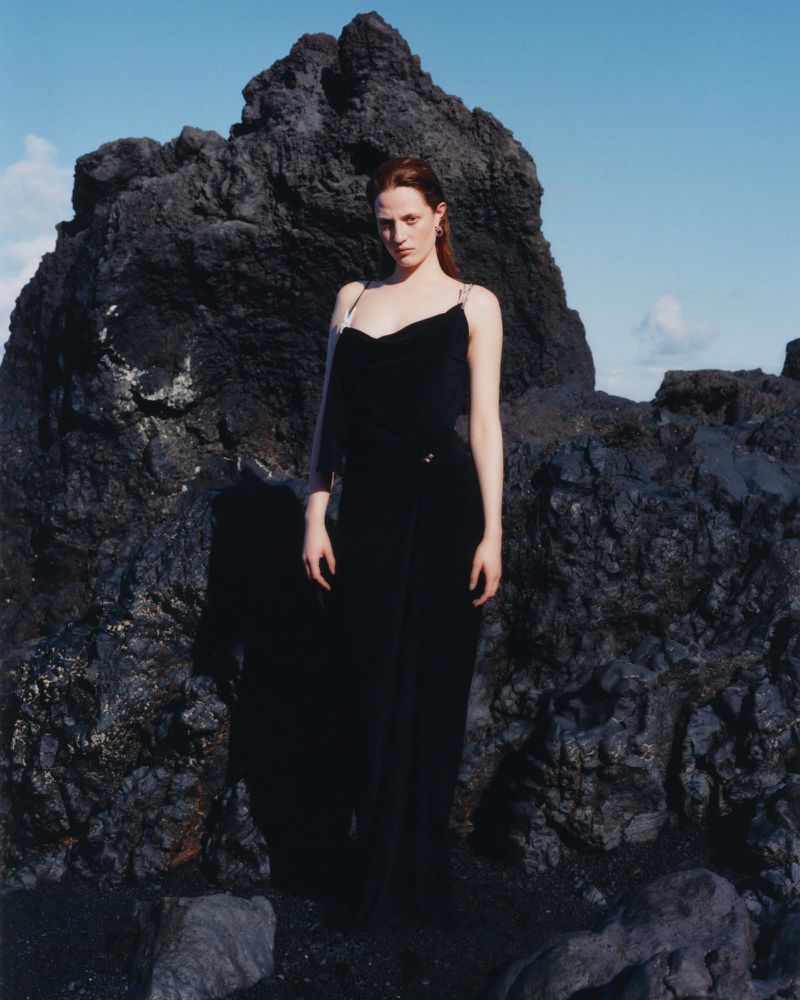 A red-haired woman in a black dress stands in front of a black rock formation under a clear sky, fashion editorial.
