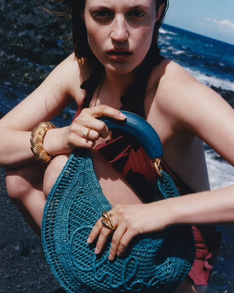 A young woman sits on a black rocky beach with the ocean in the background, holding a turquoise crochet handbag and wearing gold jewelry.