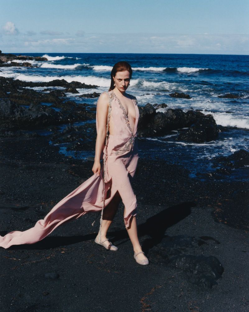 A woman in a light pink dress walks barefoot on a black sand beach with the ocean and horizon in the background on a bright, sunny day.