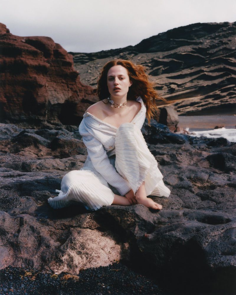 A woman with red hair sits barefoot on a rocky beach wearing a white dress, with cliffs and ocean in the background on a cloudy day.