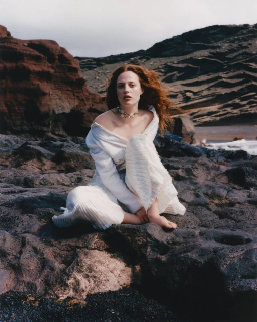 A woman with red hair sits barefoot on a rocky beach wearing a white dress, with cliffs and ocean in the background on a cloudy day.