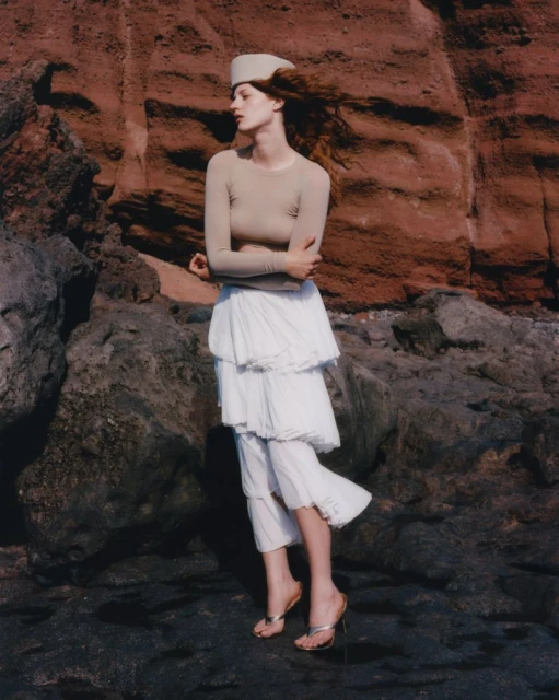 A young woman poses in an outdoor fashion shot wearing a long-sleeved beige top, tiered skirt and sandals, against a rocky beach backdrop.