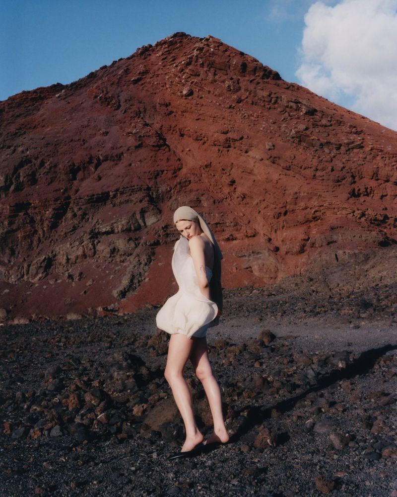 Woman in avant-garde dress poses in volcanic landscape with dark soil and a rust-colored mountain under a clear blue sky.