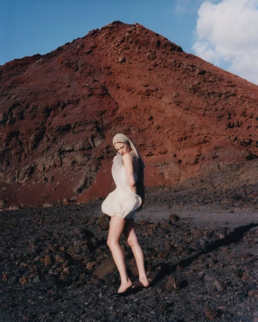 Woman in avant-garde dress poses in volcanic landscape with dark soil and a rust-colored mountain under a clear blue sky.