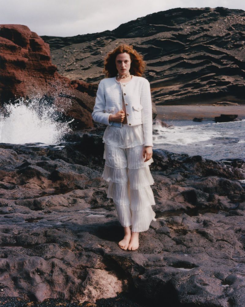 Fashion editorial image of a redhead woman in white jacket and pants on a rocky beach with breaking waves in the background.