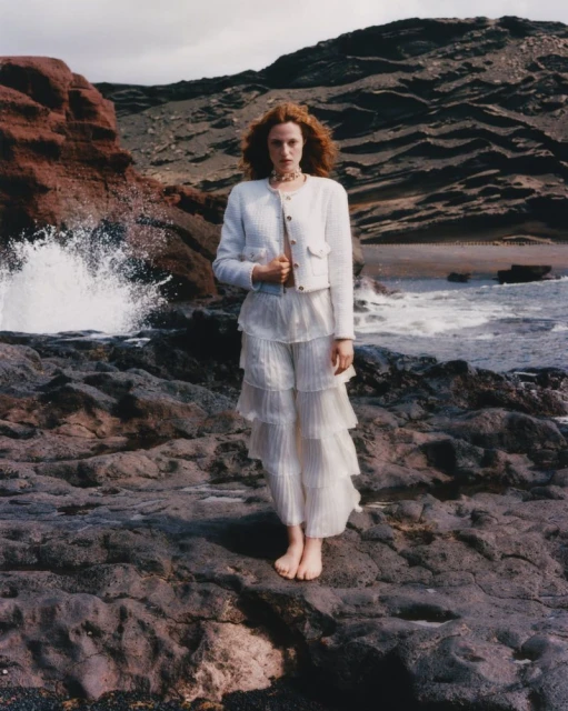 Fashion editorial image of a redhead woman in white jacket and pants on a rocky beach with breaking waves in the background.